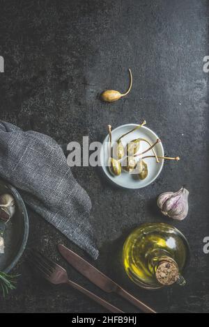 Capers on white plate at dark concrete kitchen table with olive oil, garlic, cutlery and table cloth. Mediterranean food appetizer. Pickled Italian fo Stock Photo