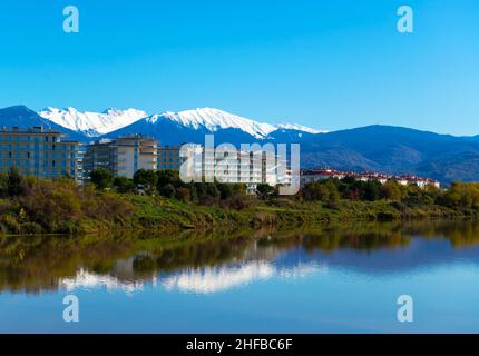 Beautiful daylight scenery of snowy Caucasus mountains with a wavy lake in front and modern buildings in the middle. Sochi, Adler Stock Photo