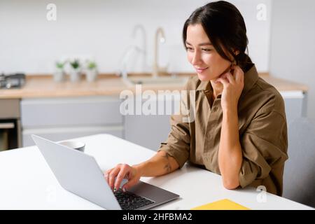 Cheerful asian girl using laptop for remote work or home leisure while sitting in the table at home. Side view of the nice girl looking at the screen with smile Stock Photo