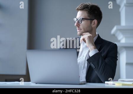 Focused young German male office worker sits at desk at home workplace distracted from laptop screen Stock Photo