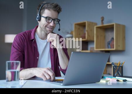 Smiling German guy sitting at home in wireless headset and taking part in online meeting Stock Photo