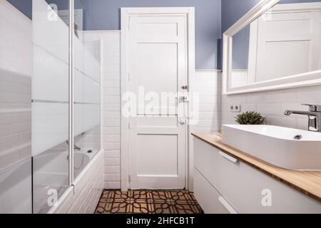 White bathroom with shower and glass partition, porcelain sink on wooden countertop and hydraulic tile floors Stock Photo