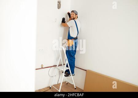 Enthusiastic electrician, handyman smiling at camera while working on installing socket on wall, standing on a ladder Stock Photo