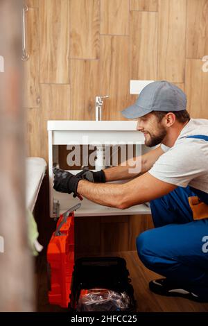 Professional plumber, male worker in uniform looking busy while fixing sink in apartment Stock Photo