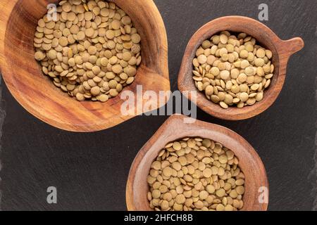 Uncooked organic green lentils in three wooden cups on a slate stone, macro, top view. Stock Photo