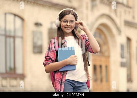 Happy kid in headphones hold school book fixing eyeglasses outdoors, audio courses Stock Photo
