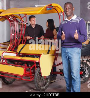 Smiling African-American pedicab driver standing near rickshaw cycle Stock Photo