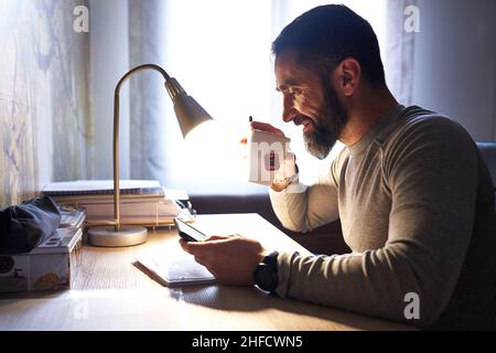 Young white man with a neat beard studying and watching the phone at his desk while taking an infusion Stock Photo