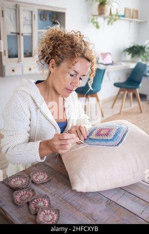 Adult caucasian woman at home doing crochet hobby activity at home decorating a shabby chic white pillow. Living room interior in background. Female p Stock Photo