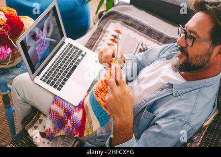 Handsome adult man learning how to knitting work with woolen yarn at home watching an online class tutorial on laptop computer. Concept of female hobb Stock Photo