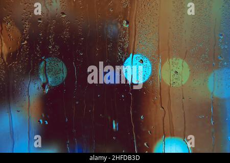 Rain drops run down the window with colorful lights of the night city. Abstract background of city life in night in rainy season. Stock Photo