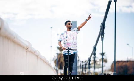 Young black man with colorful t-shirt riding an electric scooter raising his hand Stock Photo