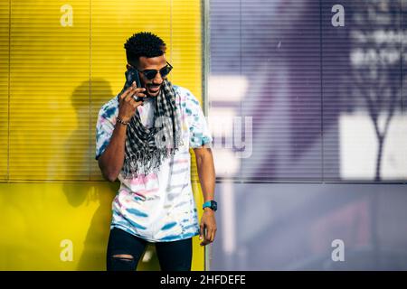 Handsome smiling black man leaning against a purple and yellow glass wall talking on his smartphone Stock Photo