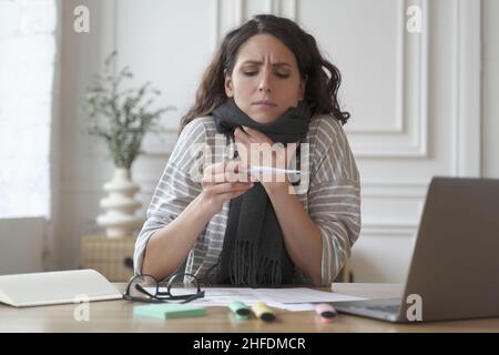 Worried young hispanic woman office worker with fever sitting workplace and looking at thermometer Stock Photo