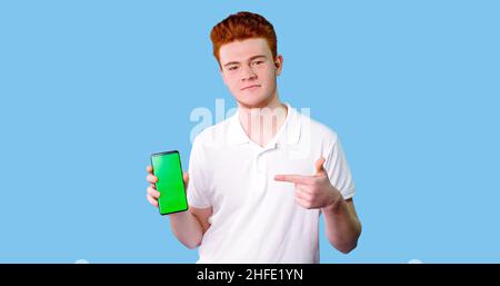 Photo of cheerful and attractive guy in white t-shirt, having smart phone with green screen in hand, pointing with forefinger to product, isolated on Stock Photo