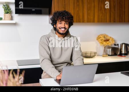 Optimistic confident dark-haired Indian handsome guy using laptop for work in the kitchen. Multiracial eastern freelancer man standing at the desk and looking at camera, smiling friendly Stock Photo