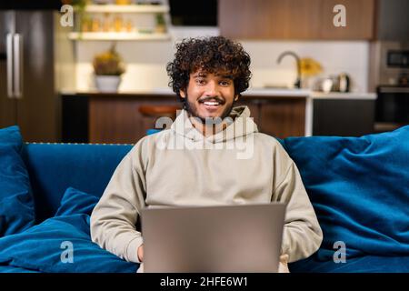 Optimistic confident dark-haired Indian handsome guy using laptop for work. Multiracial eastern freelancer man sitting at the sofa and looking at camera, smiling friendly Stock Photo