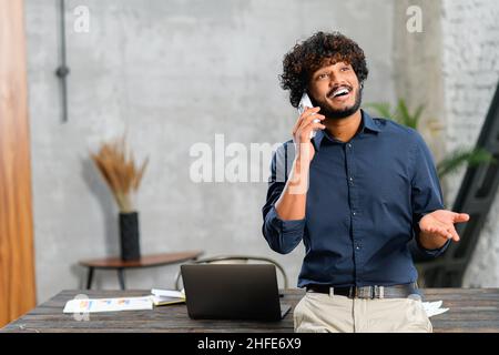 Happy middle eastern man talking on the smartphone while standing in modern loft office. Smiling successful contemporary man manager in casual wear holding phone call Stock Photo