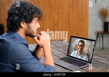 Back view of the Indian guy talking with male friend or coworker via video call. He sitting at the office desk and using laptop for video meeting. Young man on the screen Stock Photo