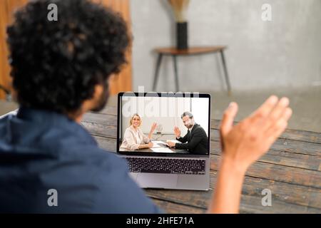 Waist up view of the young man in blue shirt waving with hand and saying hello while chatting via laptop with male and female colleagues from home Stock Photo
