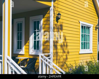 White closed windows at the yellow wooden cabin house with empty table and chairs on the terrace near the stairs in front of the room. Stock Photo