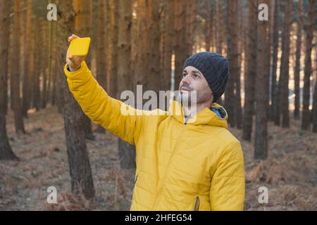 A man in a yellow coat takes a selfie with his smartphone in nature in winter Stock Photo