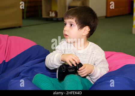 A handsome boy of 4 years old plays a game console, holds a joystick in his hands and looks away, a place for text Stock Photo