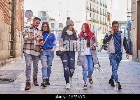 Full body happy Hispanic man in casual clothes smiling and answering phone call while walking near friends browsing cellphones, on sunny day on city street Stock Photo