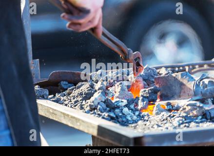 blacksmith hand holding tongs with metal piece and heating it in fire before forging . Blacksmithing concept. Stock Photo