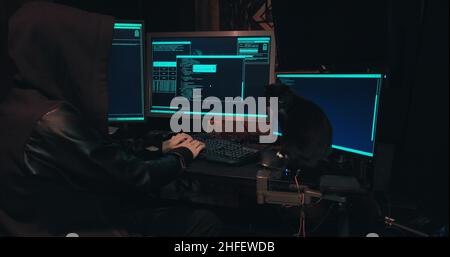 Hacker in a hood sits in front of computer screens and hacks databases Stock Photo