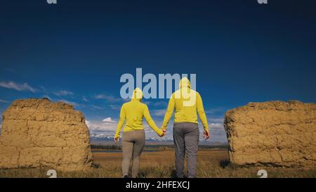 Man and woman in yellow green sportswear. Lovely couple of travelers hug and kiss near old stone enjoying highland landscape. Two travelers are walking against the backdrop of snow-capped mountains. Stock Photo