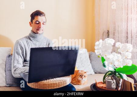 Man working online from home with pet using laptop sitting on couch. Freelancer typing on computer during covid coronavirus lockdown Stock Photo
