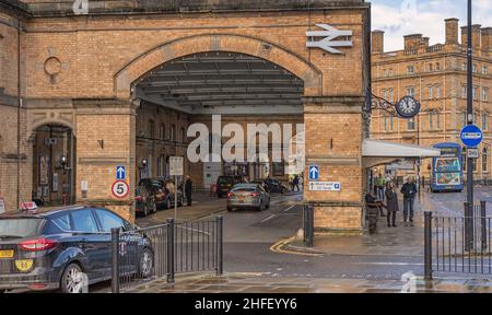 The external view of a railway station.  An arch is over the side entrance and outside people wait for a bus.  A taxi is in the foreground. Stock Photo