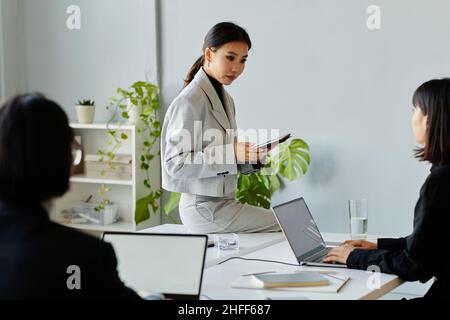 Portrait of young Asian businesswoman speaking to colleagues while sitting on table during business meeting, copy space Stock Photo