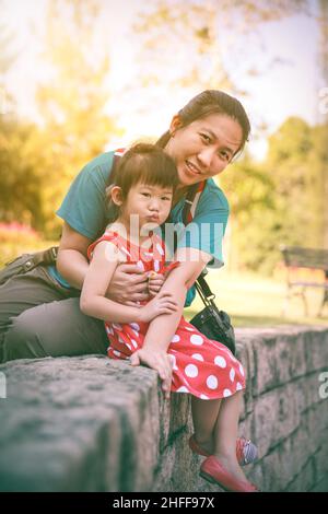 Happy asian family, mother and lovely daughter smiling and looking at camera. Playful child and parent relaxing outdoors in the day time at park with Stock Photo