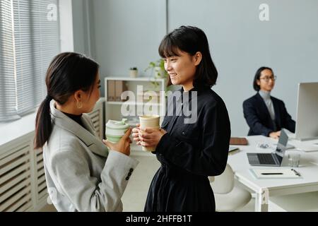 Side view portrait of two young businesswomen chatting while enjoying coffee at break in office Stock Photo