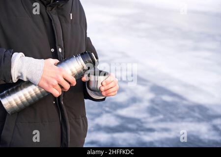 Young man, teenager pours and drinking the hot tea from thermos at the winter's beach of the cold frozen lake Baikal, people and healthy lifestyle con Stock Photo