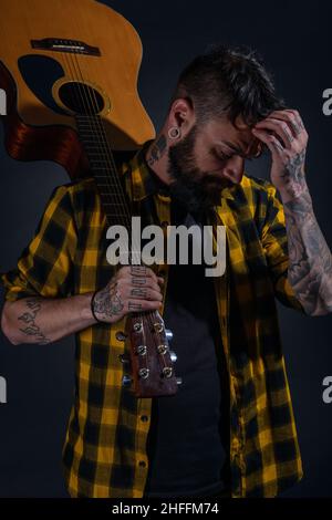 Tattooed man with a beard wearing a tartan yellow black shirt is  posing holding his guitar on his sholder Stock Photo