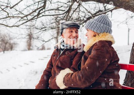Senior family couple sitting on bench outdoors during snowy winter weather. Elderly people chatting and laughing in park. Valentine's day Stock Photo