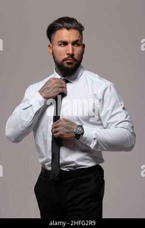 Confident Hispanic bearded man with tattooed hands in white shirt and black trousers adjusting tie and looking away while standing against gray backgr Stock Photo