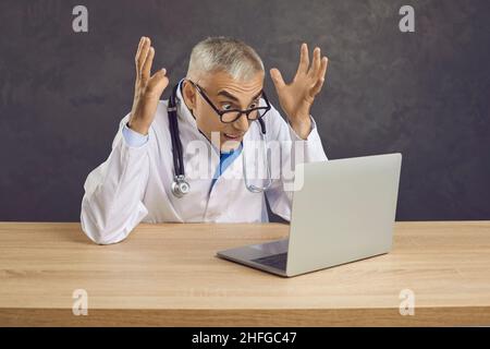 Crazy angry senior male doctor screaming in frustration looking at laptop screen. Stock Photo
