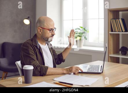 Businessman waving his hand in front of laptop webcam greeting customer on video call. Stock Photo