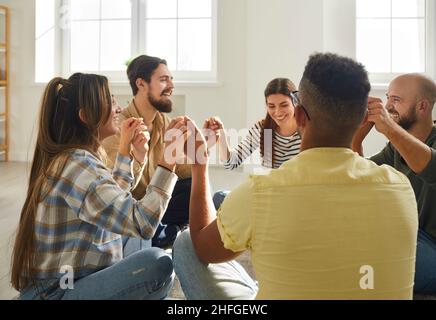 Diverse group of happy friends sitting in circle on floor, holding hands and laughing Stock Photo