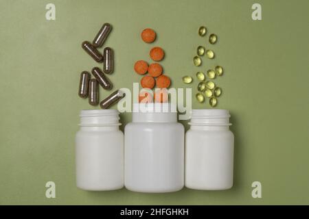 Multi-colored pills and capsuless with vitamins or dietary supplements fly out of three white plastic jars on a green background Stock Photo
