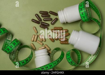 Various capsules with vitamins or dietary supplements spill out of three white plastic jars wrapped with a centimeter tape on a green background Stock Photo