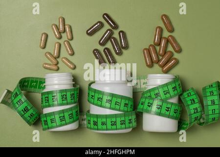 Various capsules with vitamins or dietary supplements fly out of three white plastic jars wrapped with a centimeter tape on a green background Stock Photo