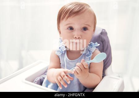 Infant baby girl in blue dress holds baby spoon and makes funny face Stock Photo