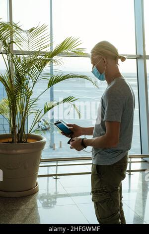 Man with power bank at the airport, charging phone to stay connected during travel, before flight at departure area. Passenger with face mask Stock Photo