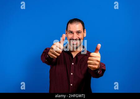 Young handsome man wearing purple shirt approving doing positive gesture with hand, thumbs up smiling and happy for success. winner gesture Stock Photo