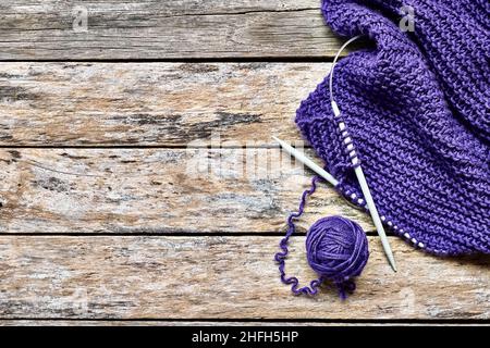 Ball of violet wool yarn, lilla scarf and wooden knitting needles on the table view from above. Hobby craft background with space for text. Concept of Stock Photo